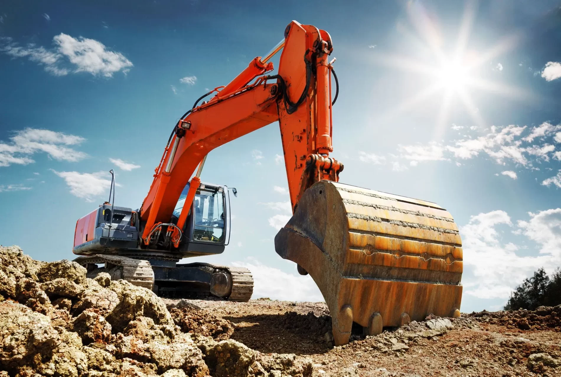 An excavator with an orange arm and large bucket rests on a construction site under a bright, sunny sky.