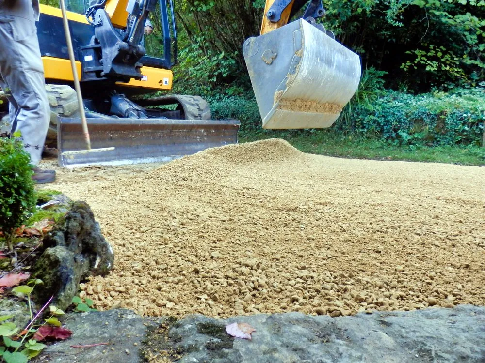 A construction vehicle spreads a layer of gravel on the ground. A person stands nearby with a rake. Green foliage is visible in the background.